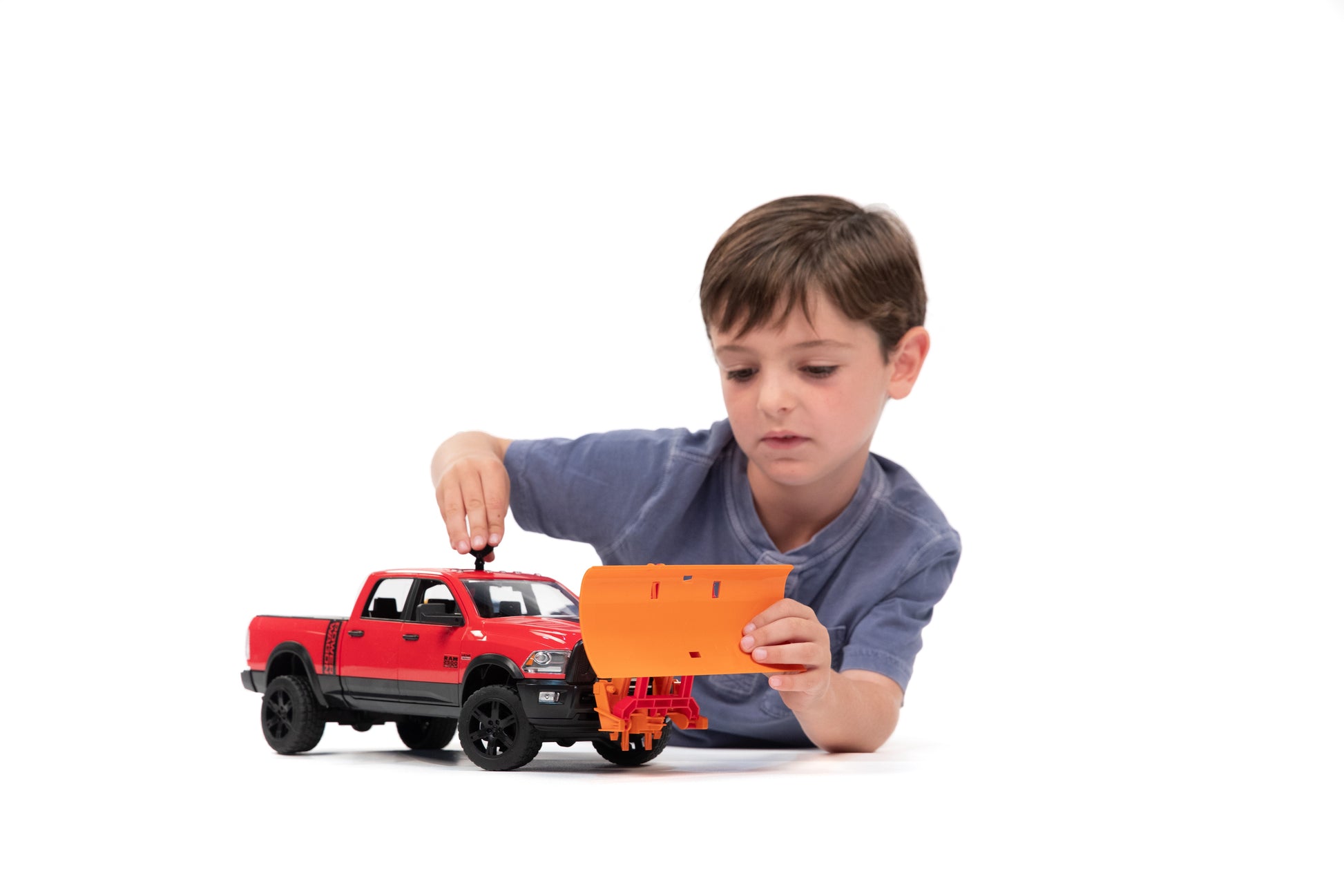 Young boy attaching bright orange Bruder Toys plow blade to red toy pickup truck for imaginative play