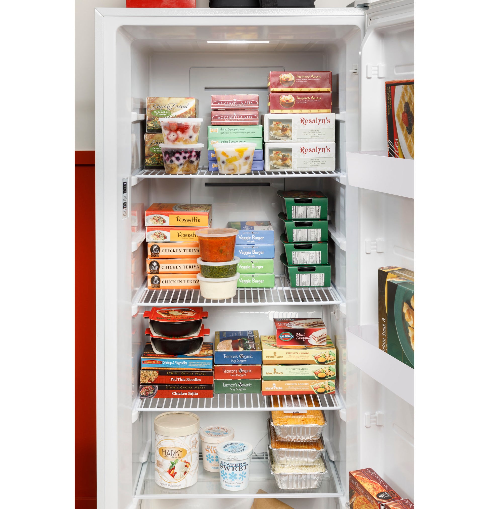 Open white upright freezer filled with organized frozen meals and desserts on wire shelves in kitchen setting