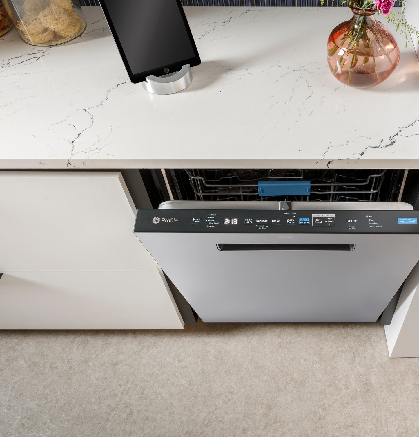 Partially open GE Profile dishwasher with stainless steel finish integrated in white kitchen cabinetry under marble countertop