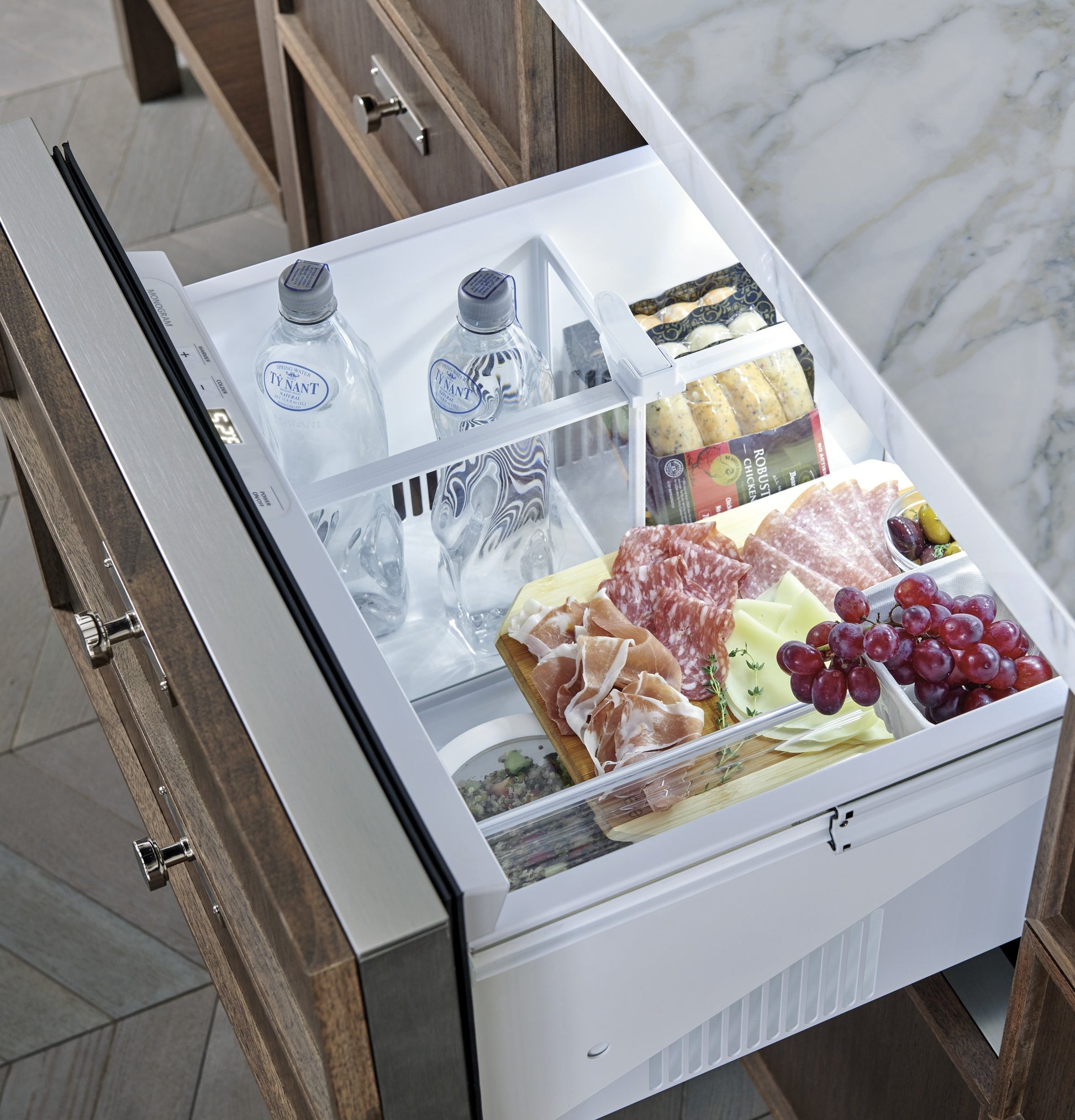 Open panel-ready double-drawer refrigerator drawer with bottled water, cheeses, meats, grapes, and crackers inside