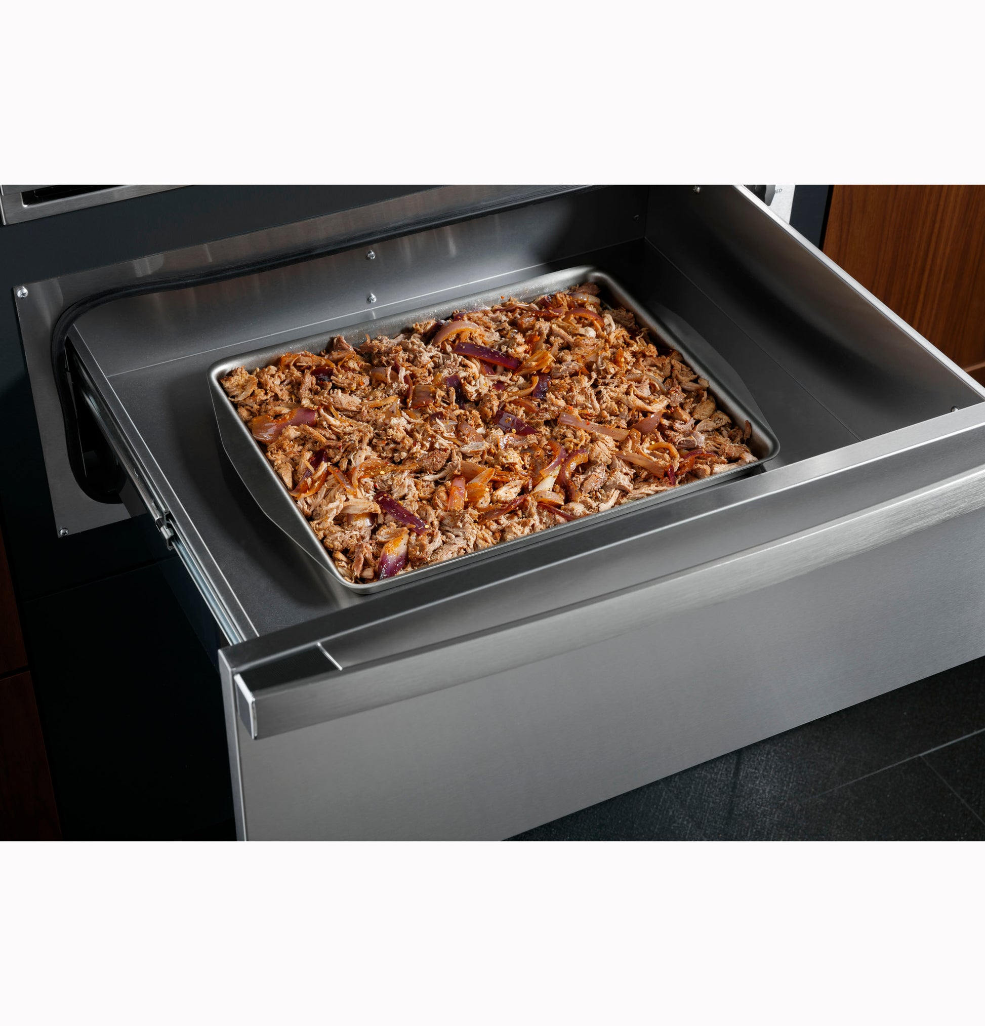 Stainless steel warming drawer open with sheet pan of cooked pulled pork in modern kitchen appliance setting