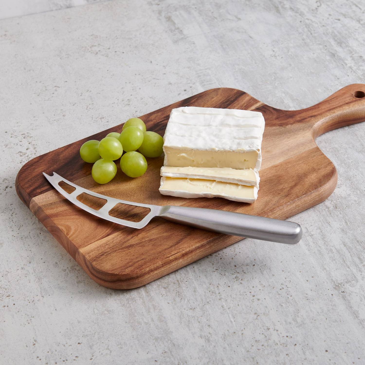 Brushed stainless steel soft cheese knife with vented blade on wooden board with Brie cheese and green grapes in kitchenware collection