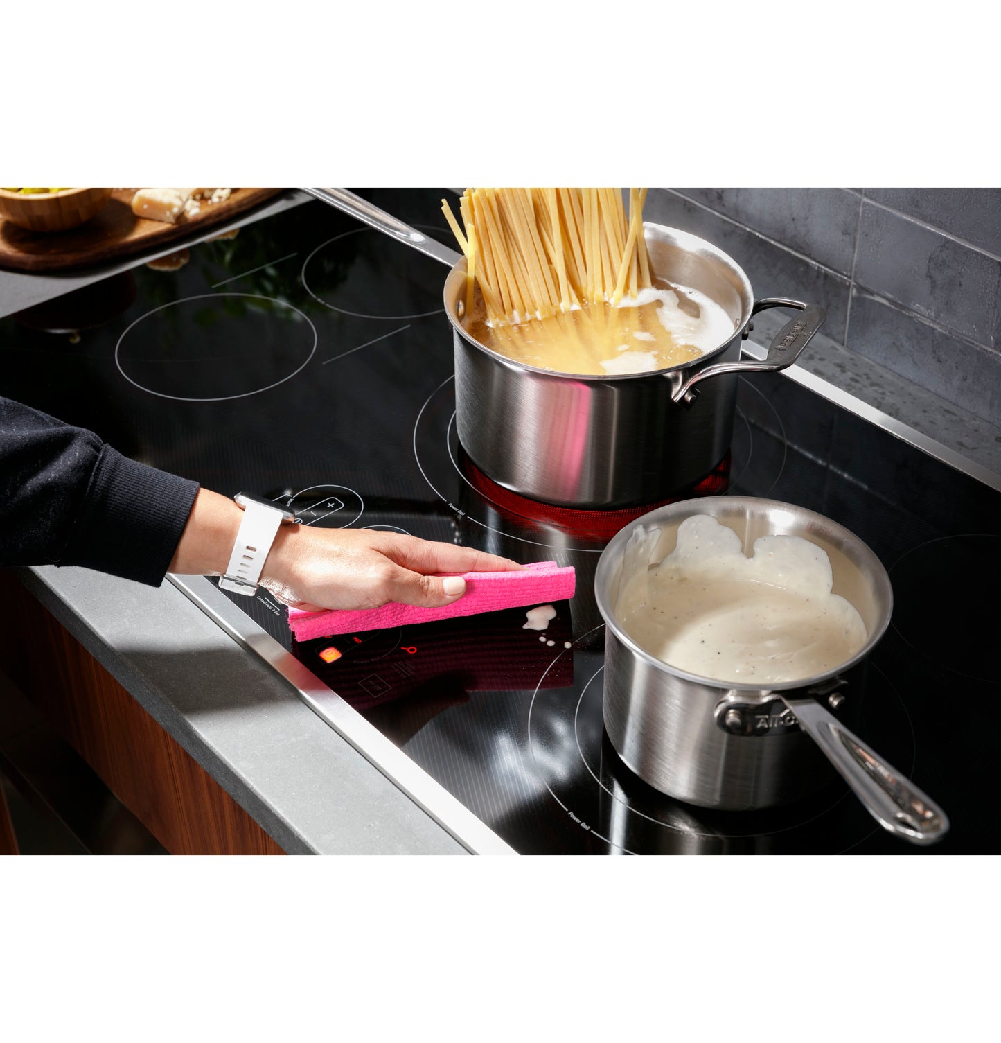 Person wiping a black touch control cooktop with spaghetti boiling in a pot and sauce in another on countertop