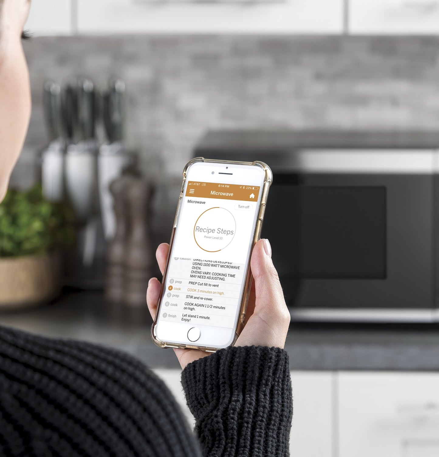 Person holding a smartphone displaying cooking instructions with a countertop microwave in the blurred background.