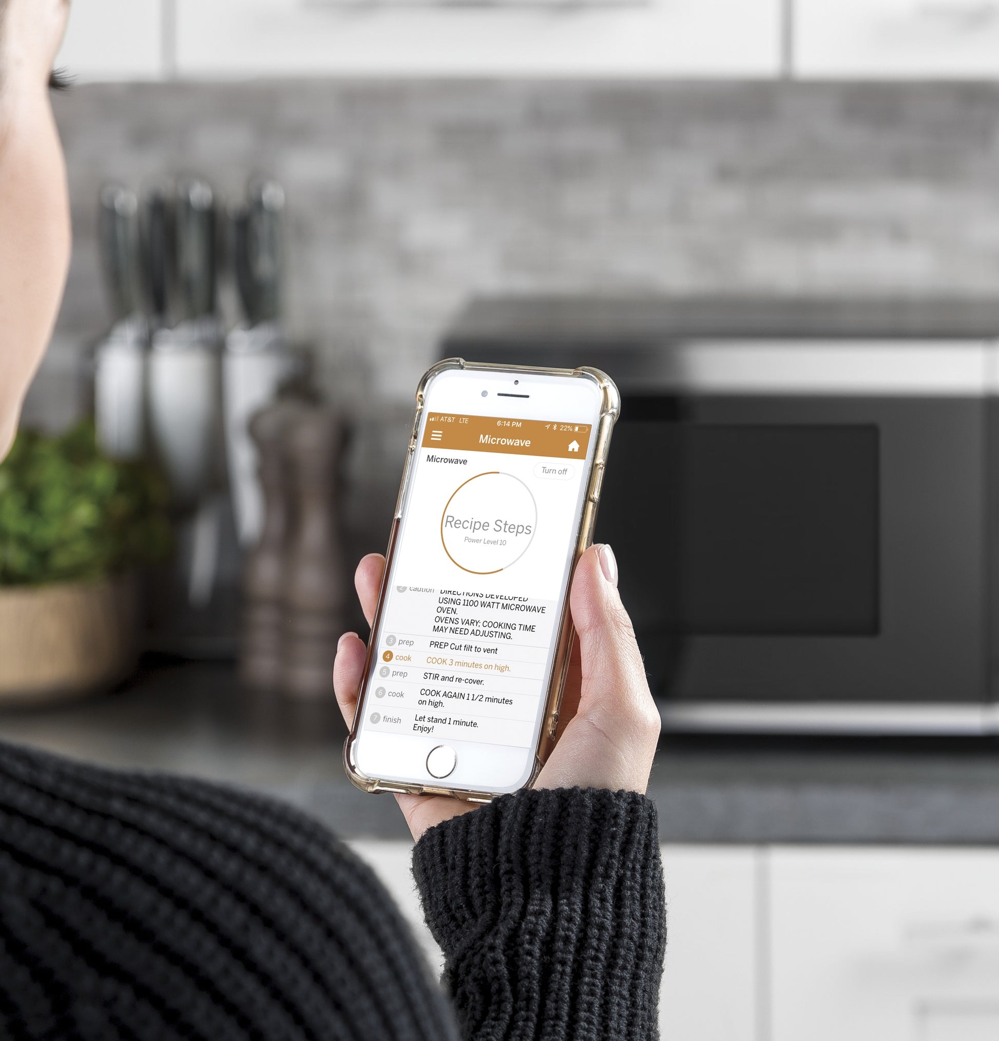 Person holding a smartphone displaying cooking instructions with a countertop microwave in the blurred background.