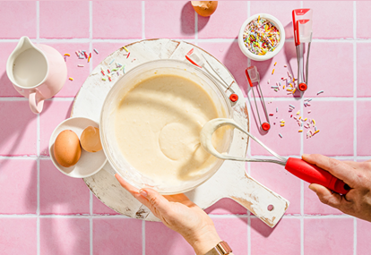 Hands using Dreamfarm Mini Flisk with red handle whisking batter on white board, surrounded by eggs and sprinkles in kitchenware setting