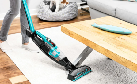 Person using Bissell ReadyClean electric blue cordless stick vacuum on rug near wooden coffee table in living room