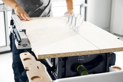 Person cutting a large wood panel on a cordless Festool table saw with dust extraction in a workshop setting