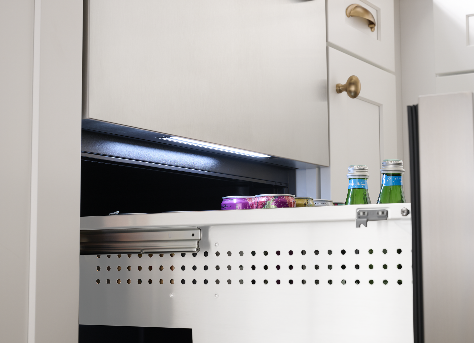 Under-counter stainless steel fridge drawer open with drinks and snacks stored in white kitchen cabinetry