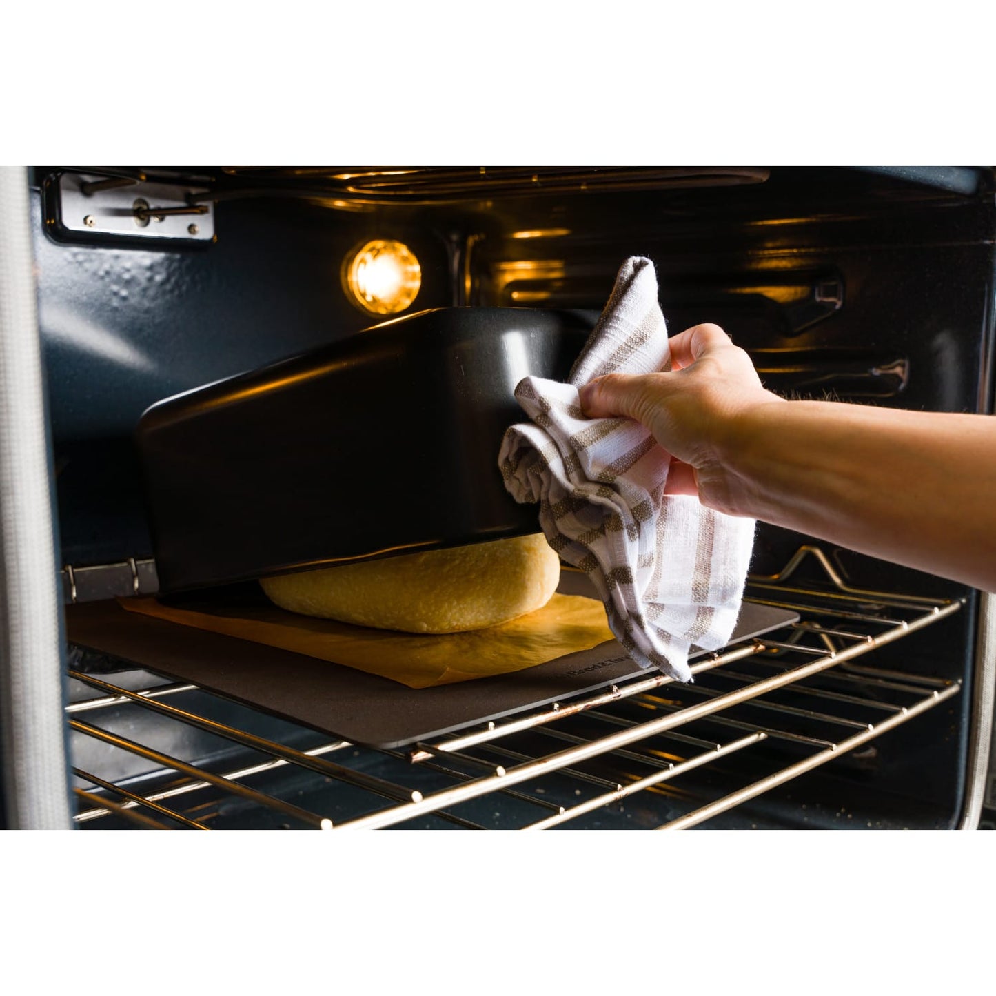 Hand placing a matte black baking shell over dough inside an oven for kitchenware and bakeware collections under $100