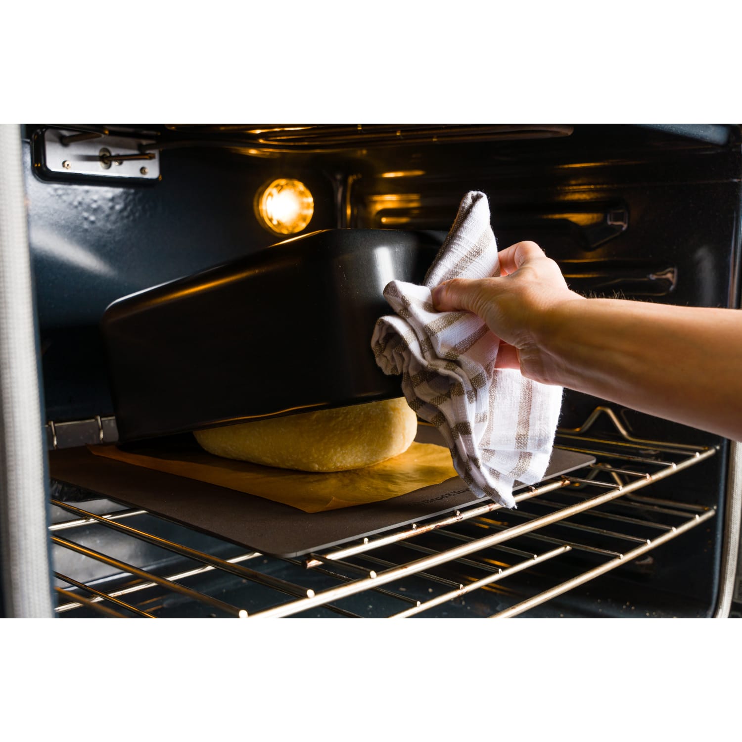 Hand placing a matte black baking shell over dough inside an oven for kitchenware and bakeware collections under $100