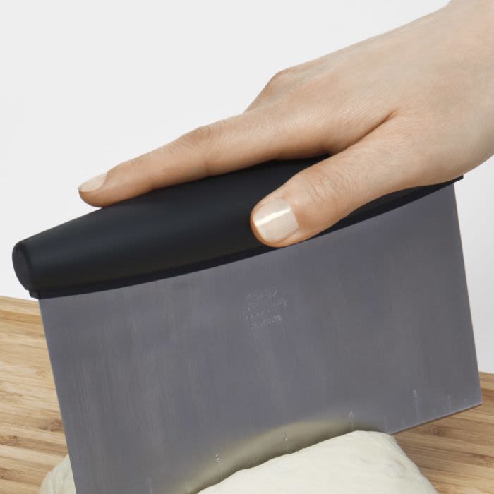 Hand holding black-handled stainless steel scraper cutting dough on wooden surface in kitchenware setting