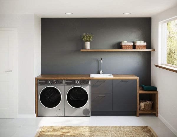 Modern laundry room with Bosch compact washer and dryer in pearl steel, dark cabinetry, and wooden countertop with shelf above