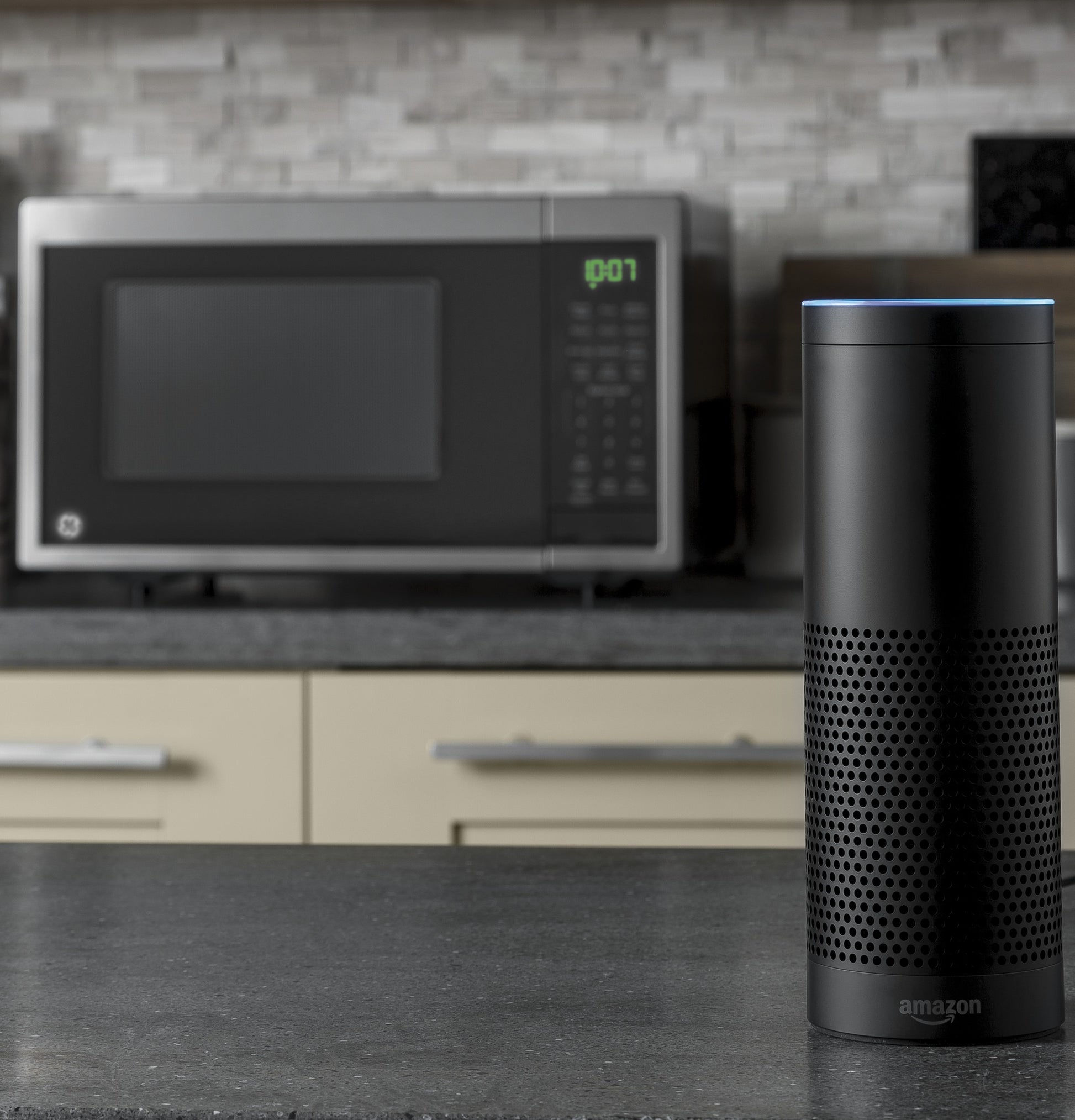 Black smart speaker with blue light ring on countertop and stainless steel microwave in background in kitchen setting