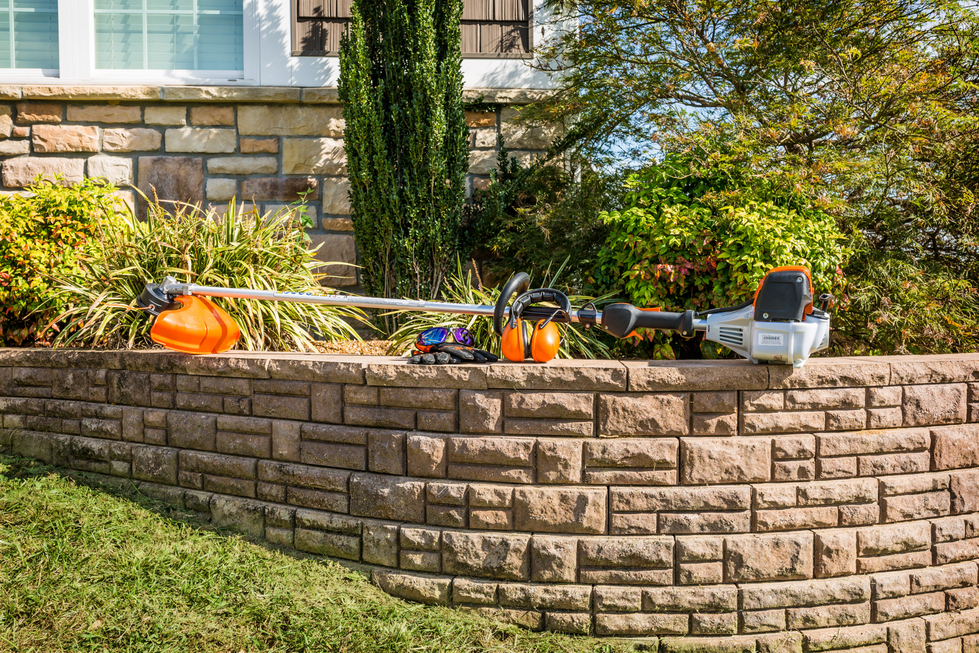 Orange and silver straight shaft string trimmer resting on retaining wall for home improvement and landscaping tasks
