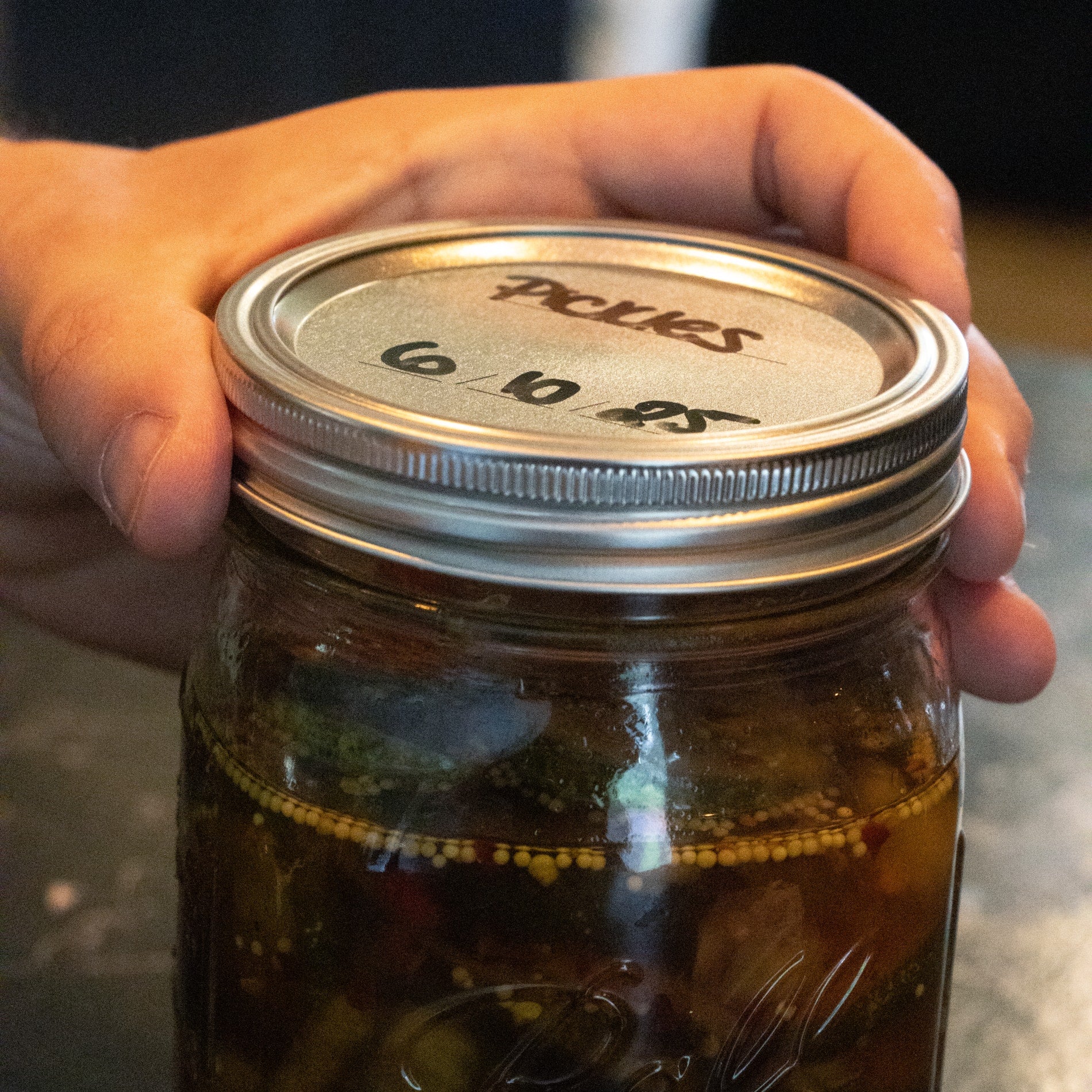 Hand holding a glass wide-mouth jar sealed with a silver metal lid labeled with handwritten contents and date, suitable for Kitchenware and Under $50 collections