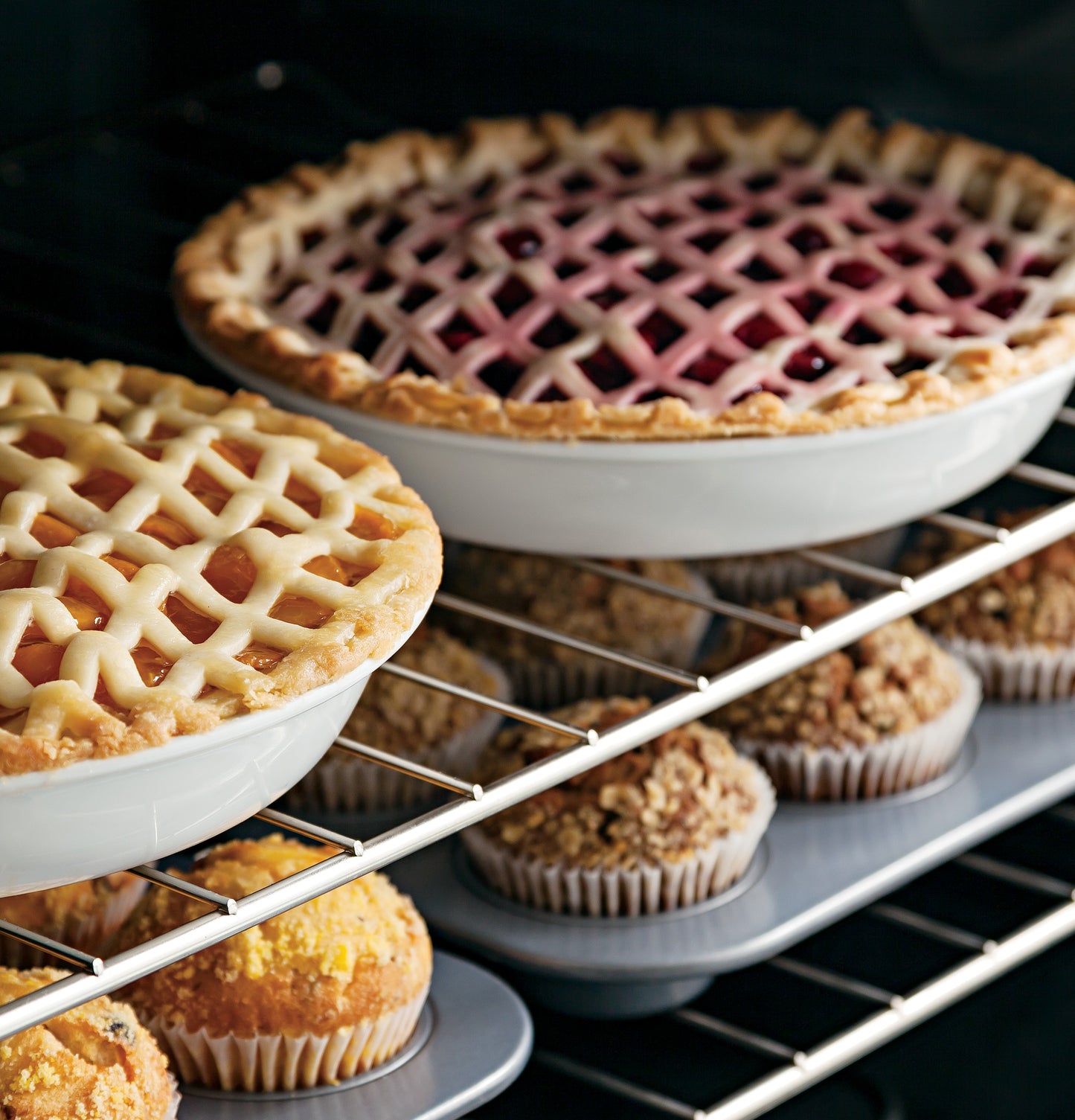 Two lattice-topped fruit pies and muffins baking together inside a free-standing kitchen range oven rack