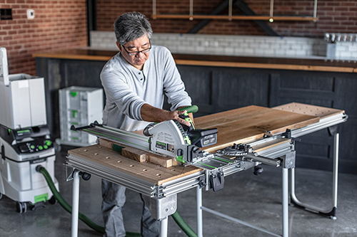 Man using Festool cordless track saw with guide rail cutting wood on portable workbench in workshop setting