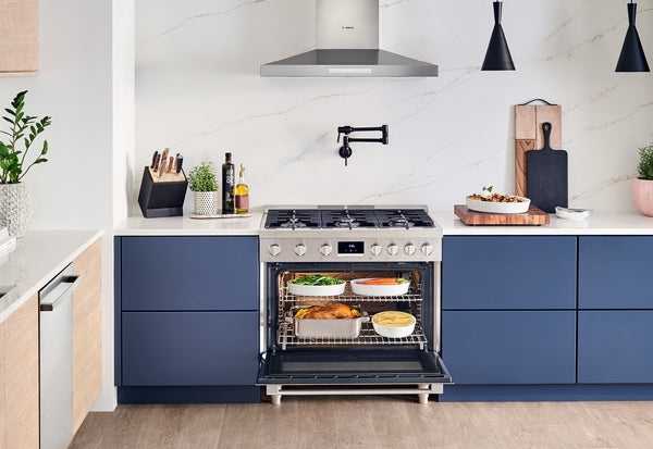 Modern kitchen with stainless steel range hood, blue cabinetry, and oven baking multiple dishes on wooden floor
