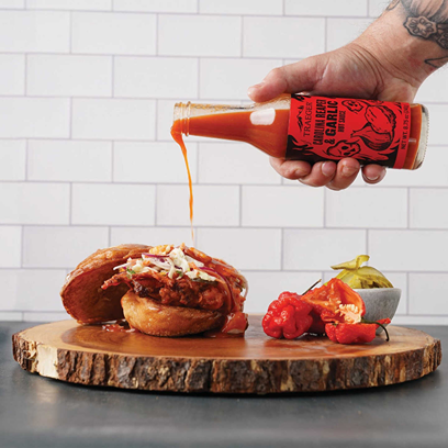 Hand pouring Traeger Carolina Reaper and garlic hot barbeque sauce over sandwich on wooden serving board