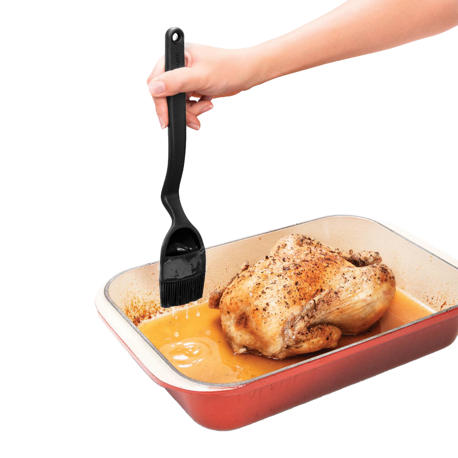 Hand holding black Brizzle Basting Brush with curved handle basting roasted chicken in red roasting pan for kitchen utensils