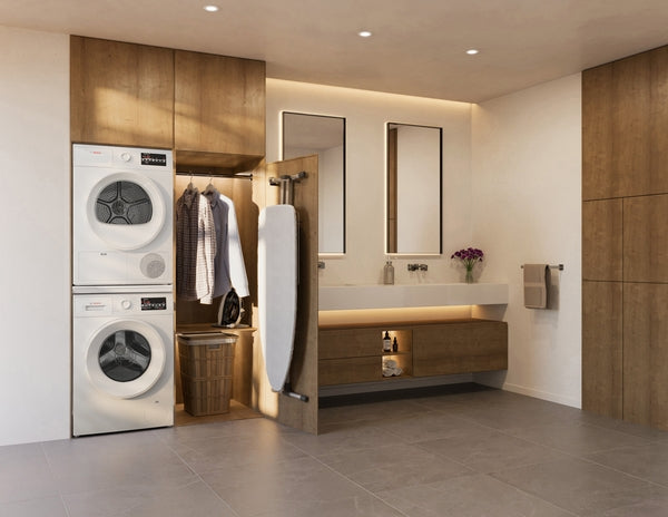 Modern laundry area with stacked white compact Bosch condensation dryer and washer beside wooden cabinets and ironing board