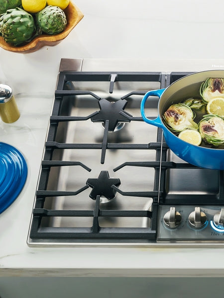 Stainless steel gas cooktop with star burner and simmer burner, blue pot cooking vegetables surrounded by kitchenware and artichokes