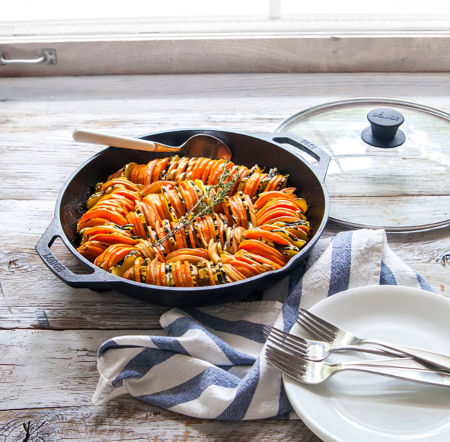 Cast iron skillet with sliced seasoned vegetables arranged in rows beside white plates and utensils on rustic wood table