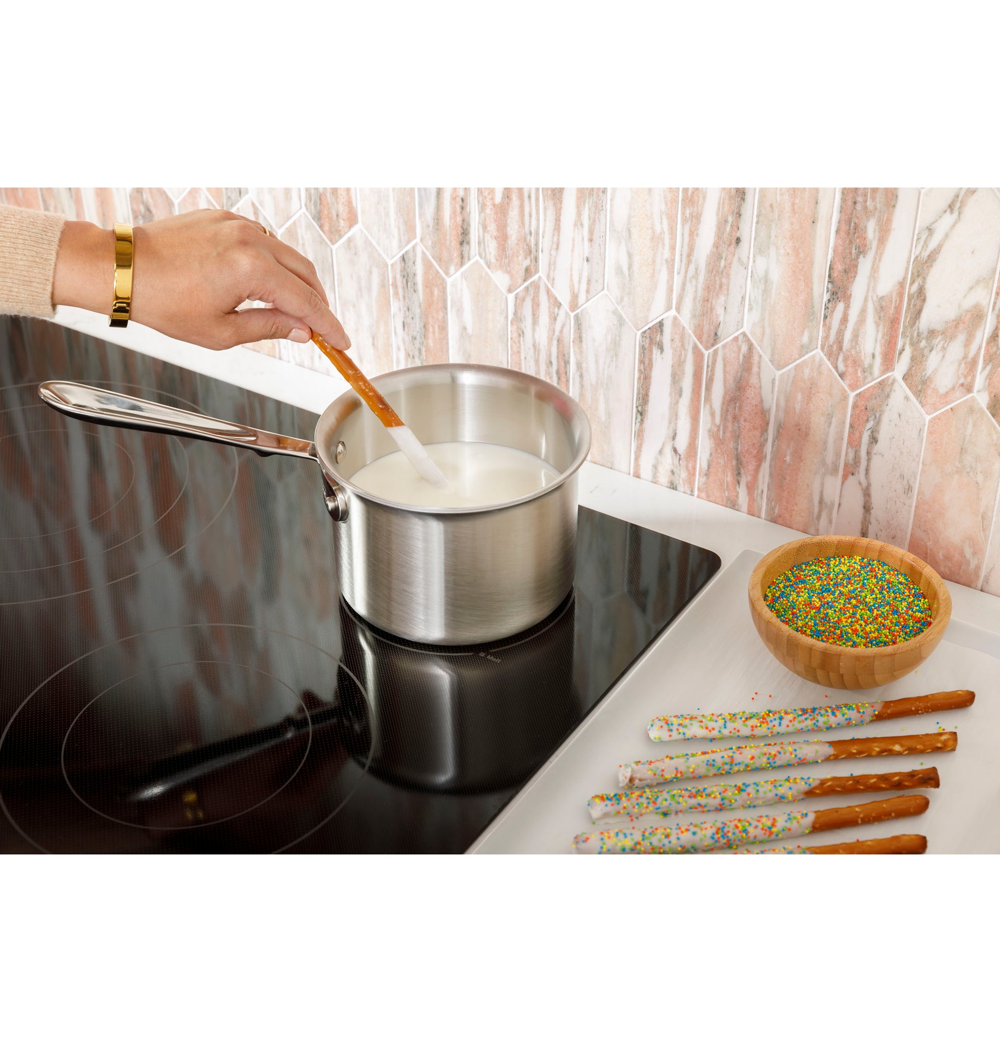 Person dipping a sprinkle-covered pretzel into milk heated in a stainless steel pot on a black electric cooktop surface