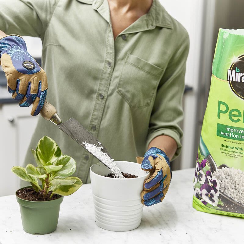 Person wearing gloves scooping perlite into white planter near green potted plant and Miracle Gro bag
