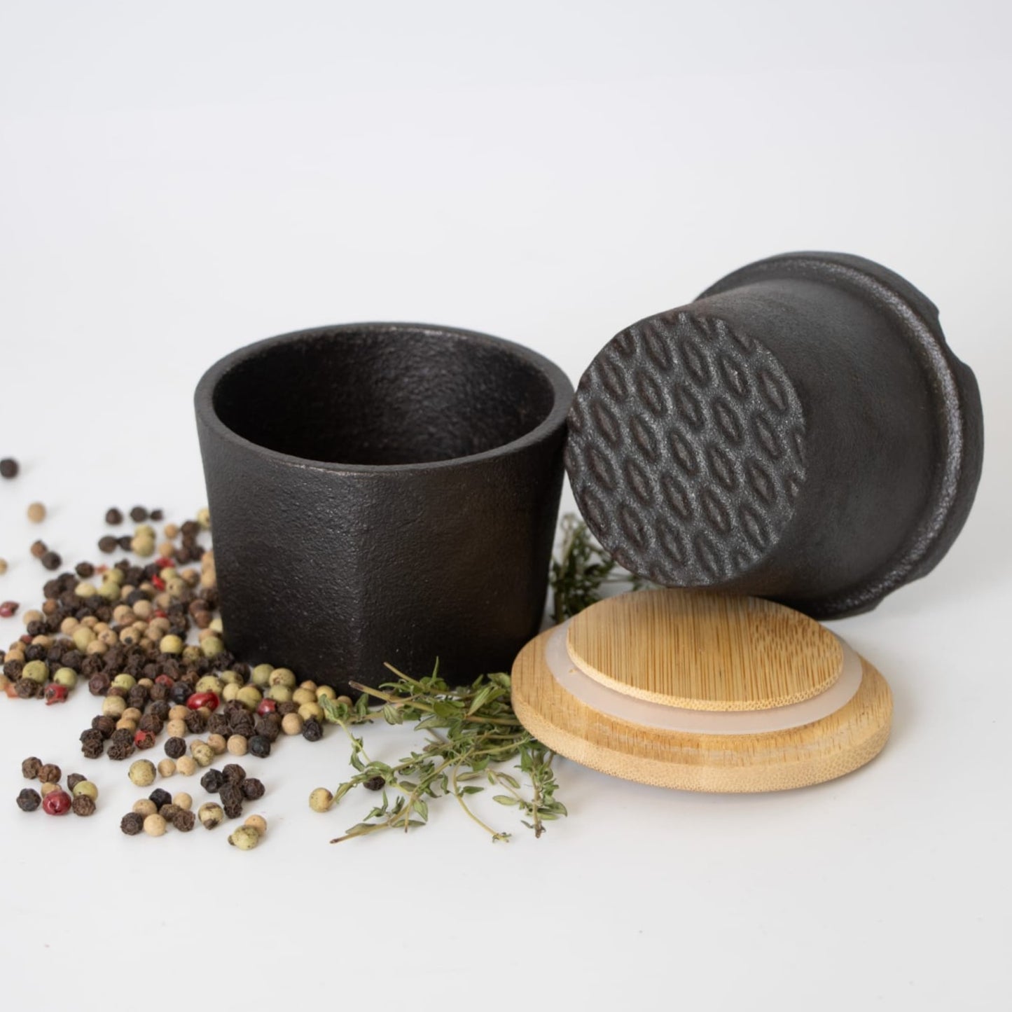 Set of cast iron spice grinders with bamboo lids surrounded by mixed peppercorns and fresh herbs in kitchen gadgets collection