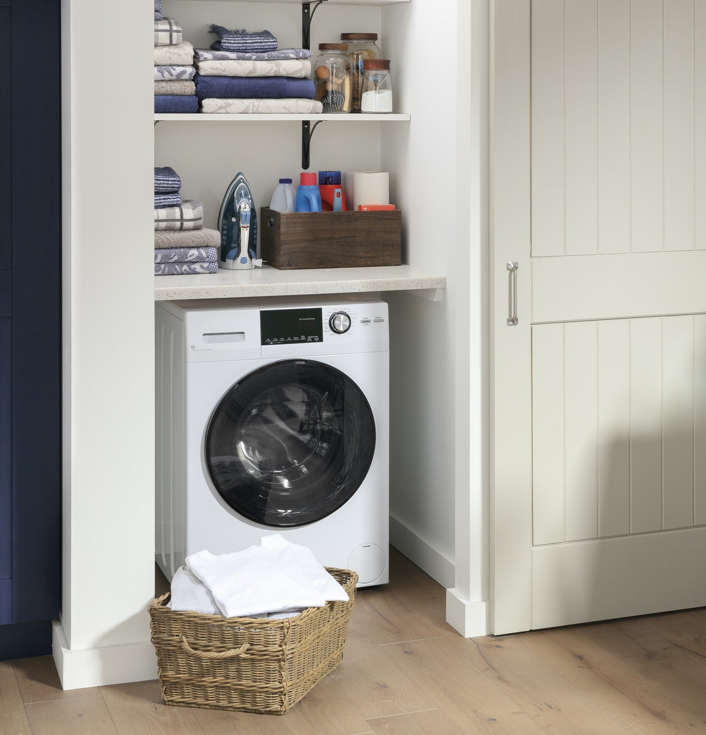 Compact white front load washer installed under countertop in laundry nook with organized shelves and laundry basket nearby