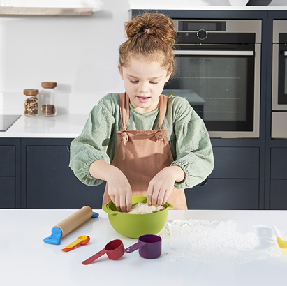 Child wearing apron mixing dough in green bowl with colorful measuring spoons and rolling pin in modern kitchen setting