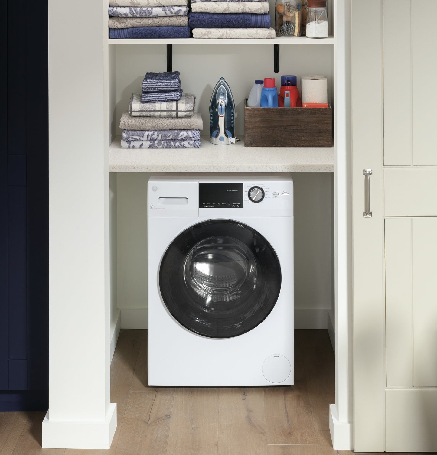 White front load washer/dryer combo installed in a built-in laundry nook with folded towels and laundry supplies above