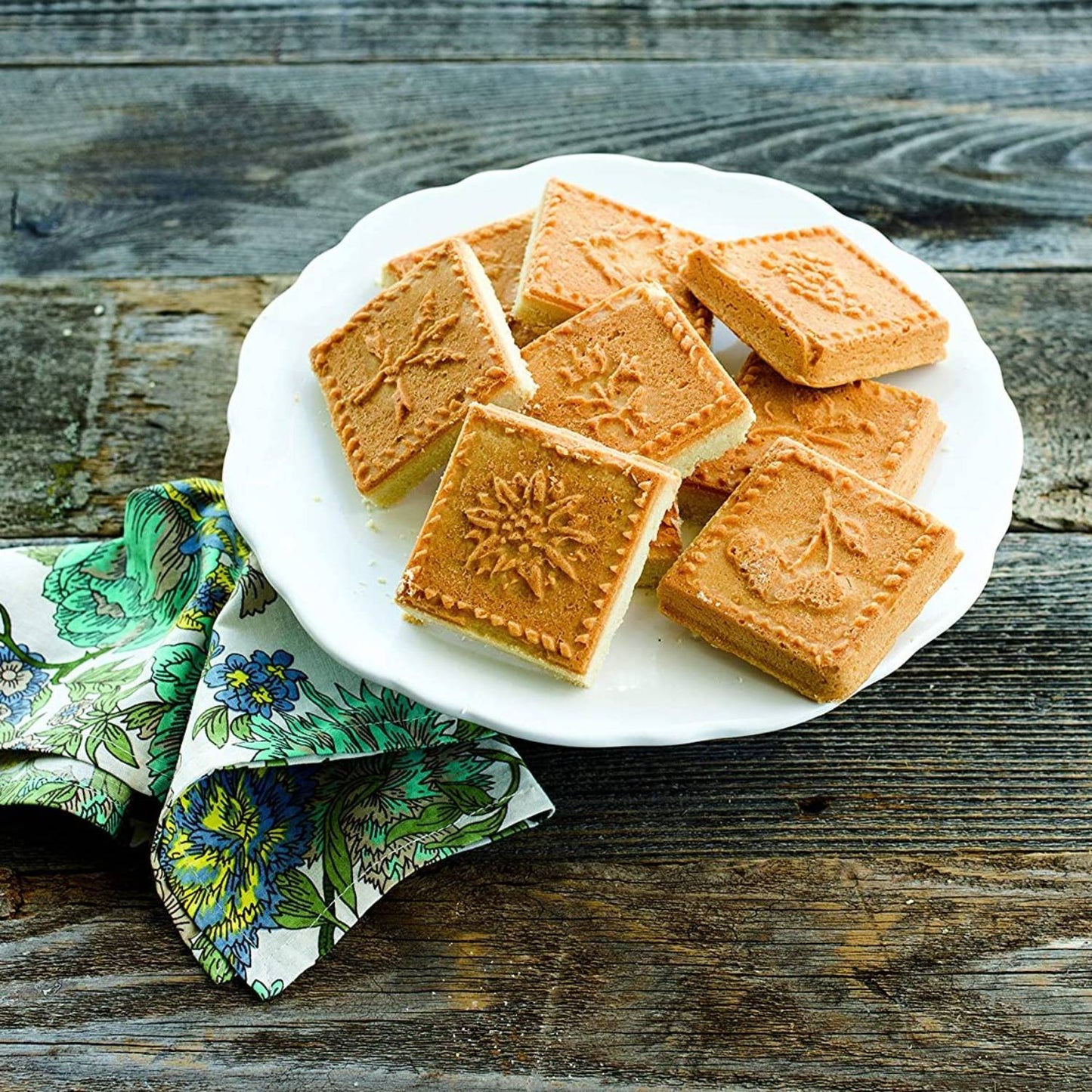 Plate of square shortbread cookies with embossed floral and nature designs on rustic wooden table with floral napkin