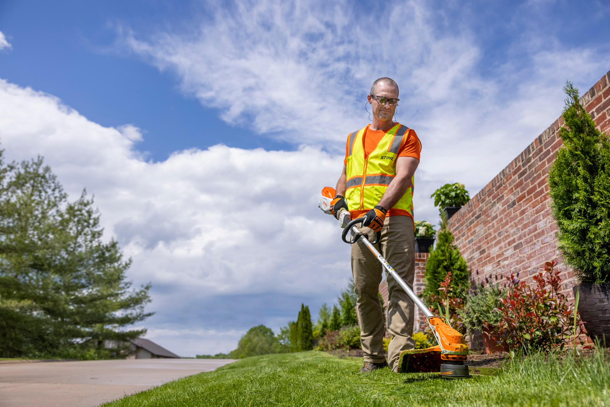 Person wearing safety vest using orange and white battery-powered trimmer for outdoor lawn and home improvement tasks