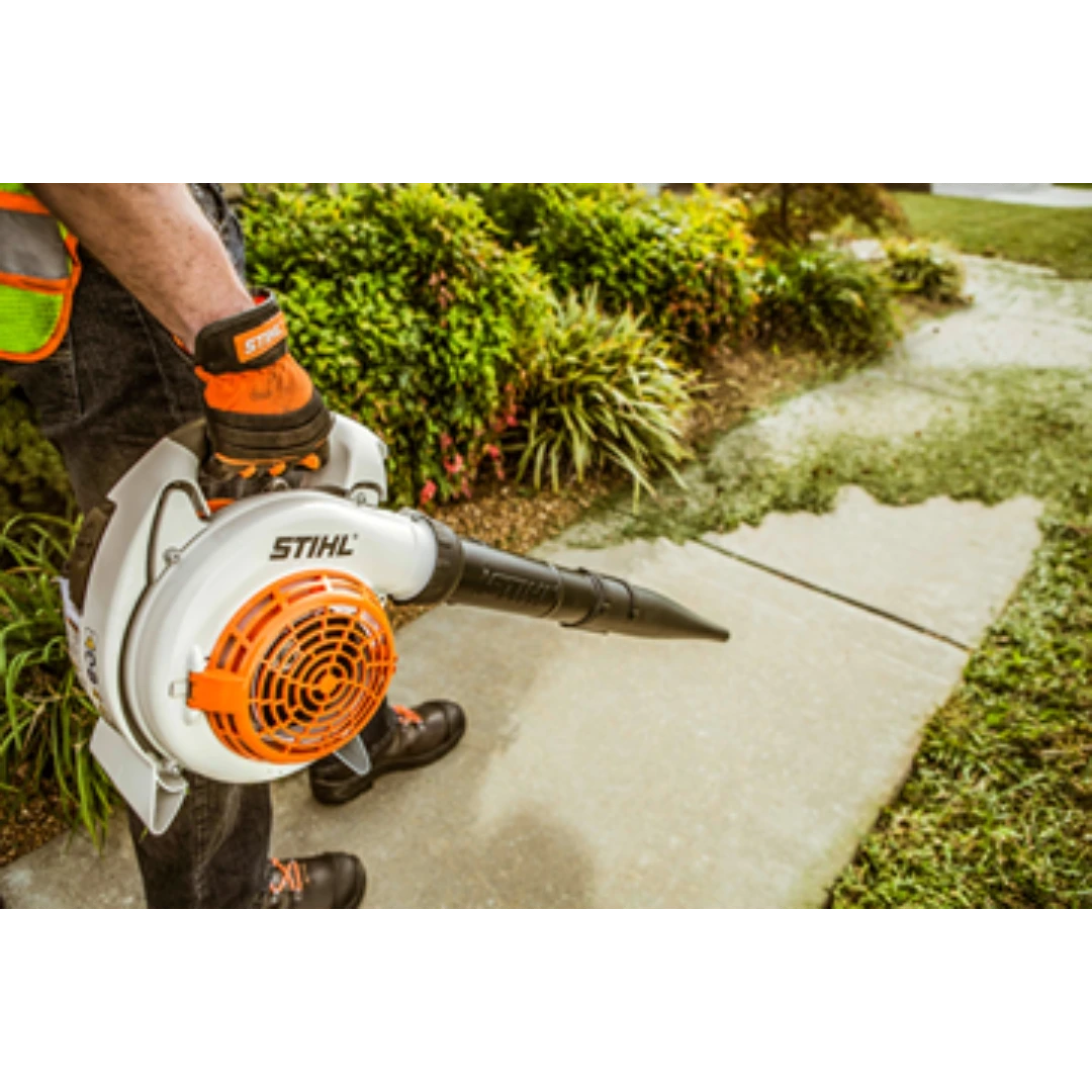 Person using a gas-powered handheld blower with white and orange housing to clear leaves from a sidewalk in a garden setting