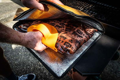 Person placing insulated Drip EZ Rest EZ BBQ Blanket on grill shelf for heat retention during cooking