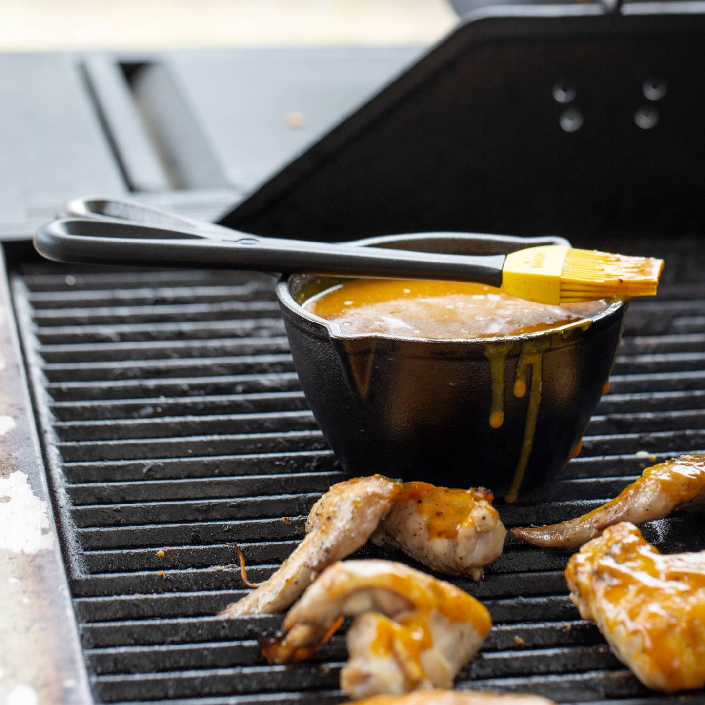 Black cast iron melting pot holding sauce with yellow silicone brush resting on grill beside cooking chicken wings