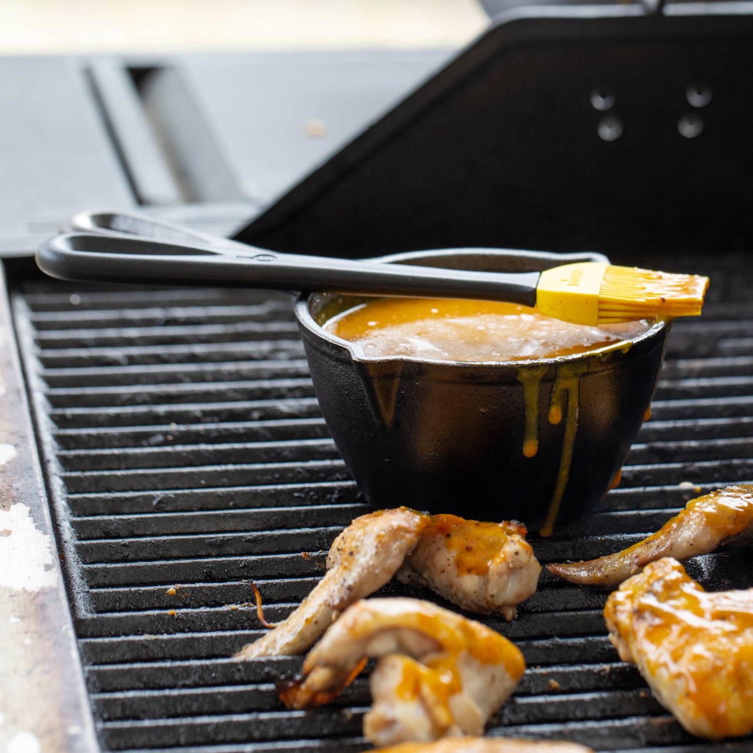 Black cast iron melting pot holding sauce with yellow silicone brush resting on grill beside cooking chicken wings