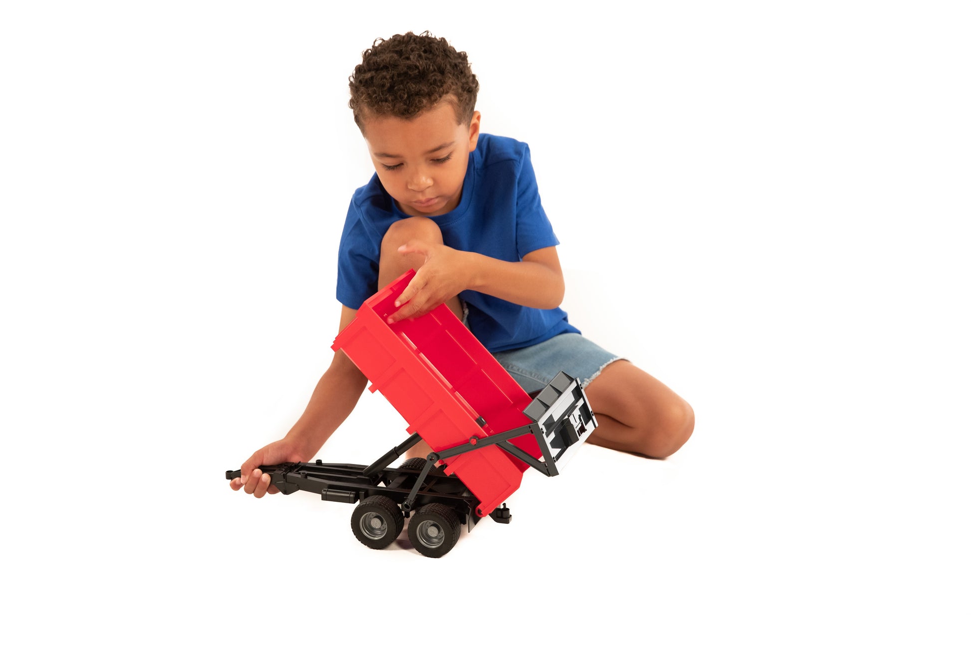 Child in blue shirt playing with a red tipping trailer toy featuring black frame and dual wheels on white background