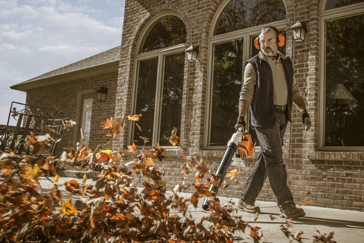 Man wearing ear protection using a gas-powered blower to clear fallen leaves from a walkway outside a brick home
