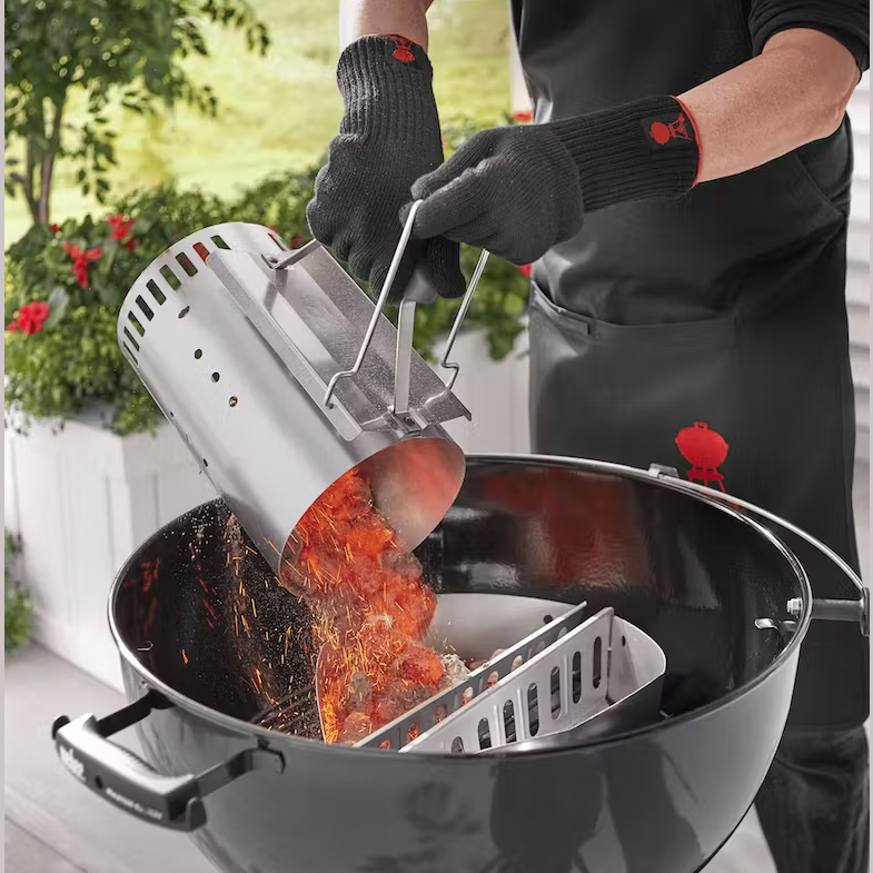 Hands wearing black heat-resistant gloves pouring flaming charcoal briquettes from steel chimney starter into kettle grill outdoors