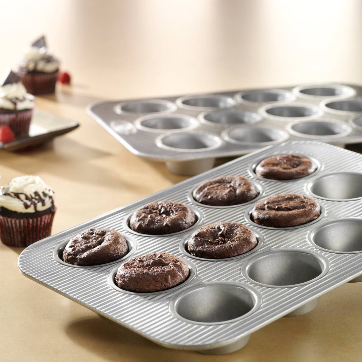 Textured metal muffin pan with twelve cups, partially filled with freshly baked chocolate muffins on wooden table