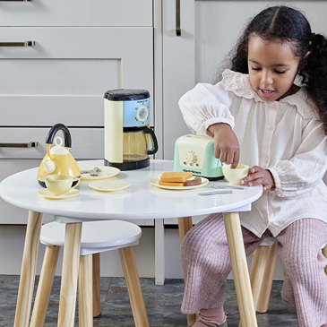 Child sitting at wooden table playing with pastel-colored toy kitchen appliances and breakfast accessories
