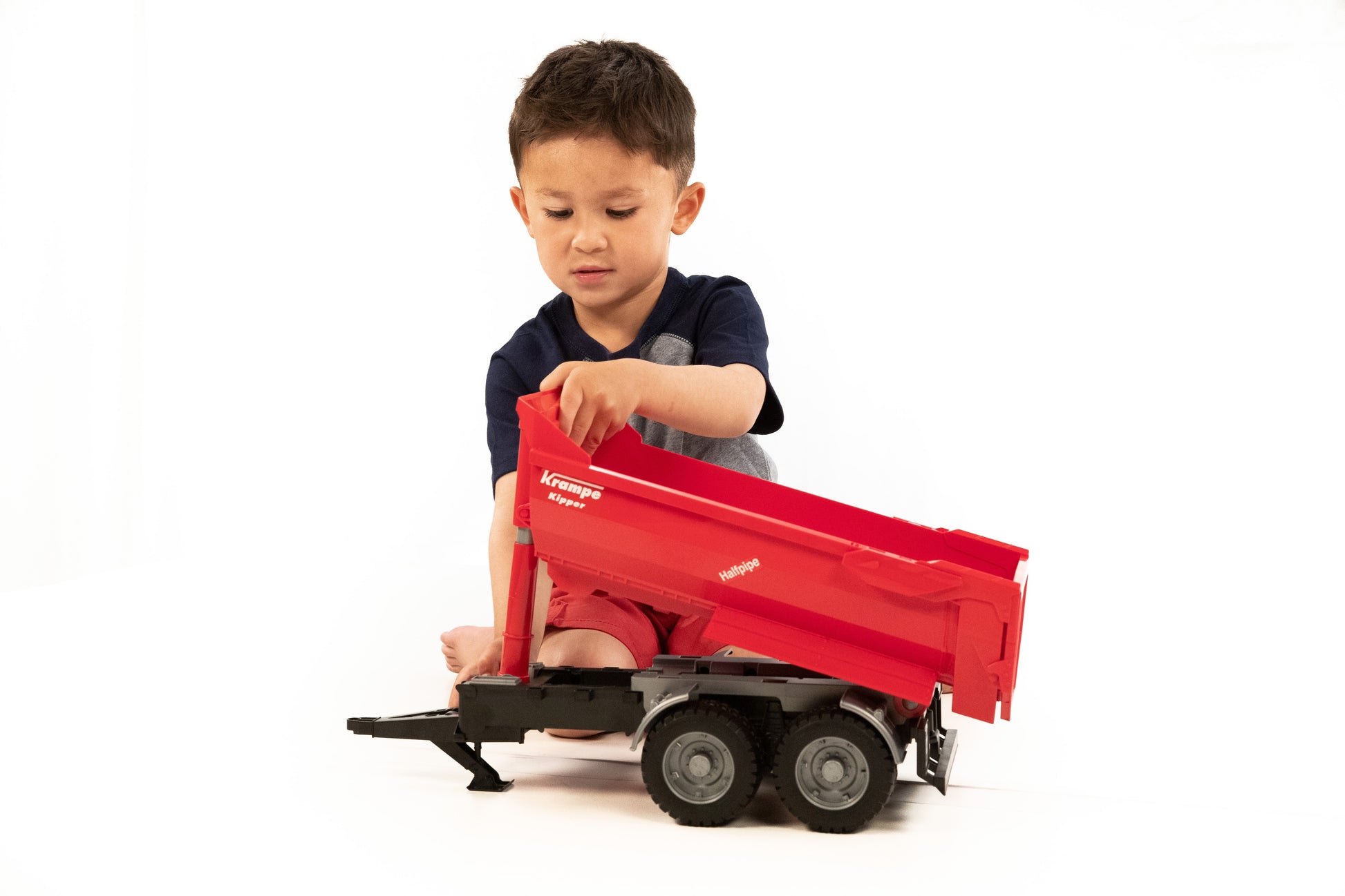 Child playing with red tandem halfpipe tipping trailer toy with black chassis and detailed wheels on white background