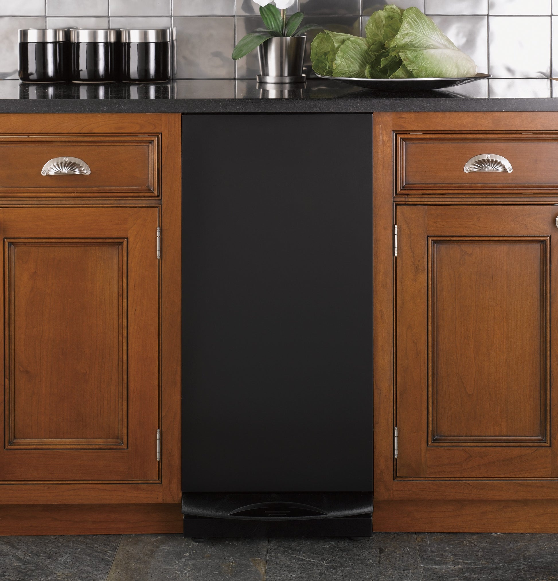 Matte black built-in compactor installed between wooden kitchen cabinets with dark countertop and backsplash tiles