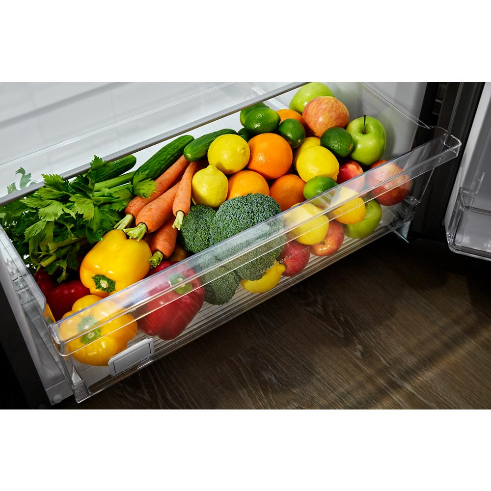 Clear refrigerator drawer filled with fresh assorted vegetables and fruits on wooden kitchen floor