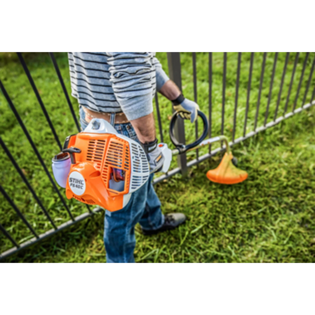 Person holding orange and white gas-powered curved shaft trimmer near metal fence on green grass