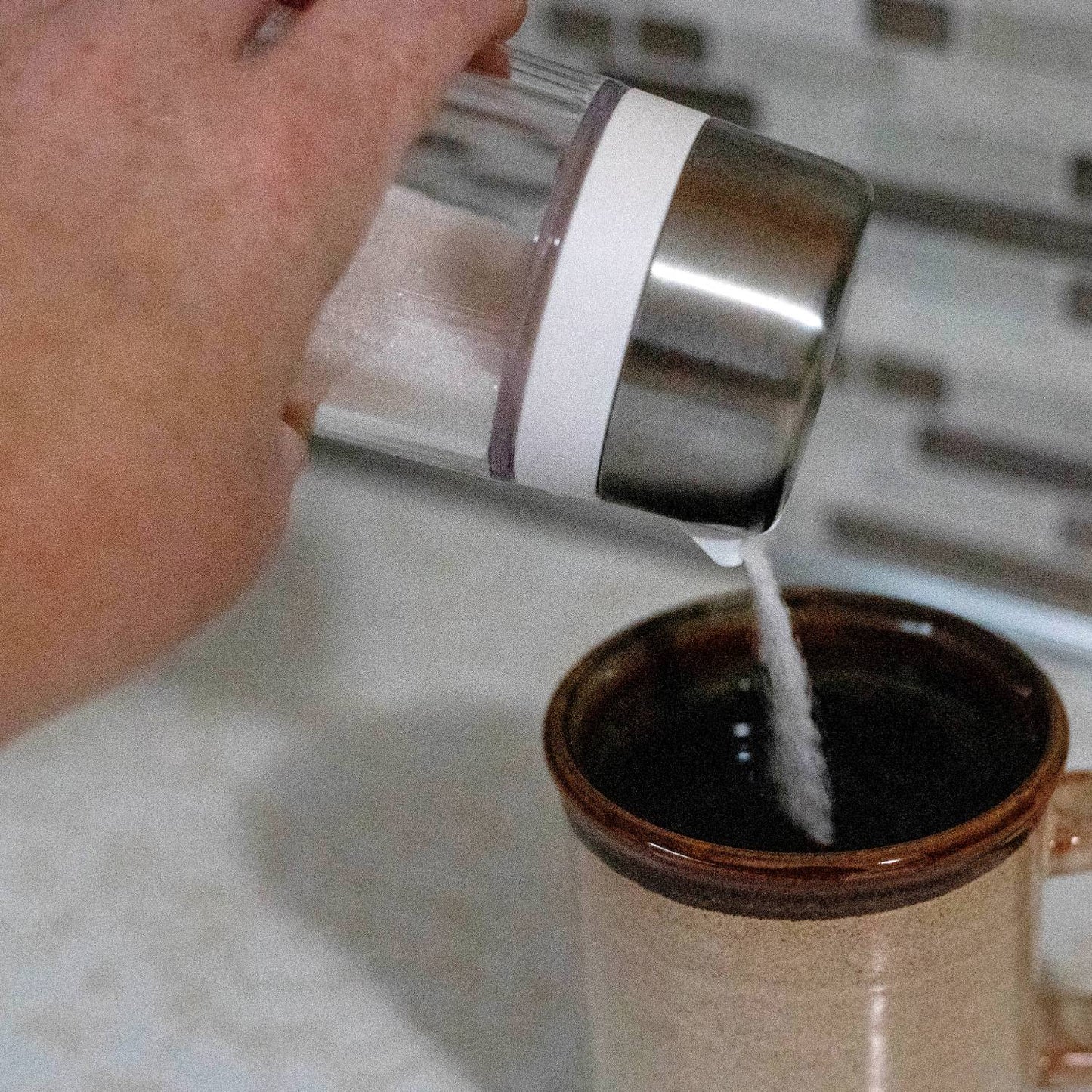 Hand pouring sugar from clear dispenser with stainless steel lid into brown ceramic mug on kitchen countertop